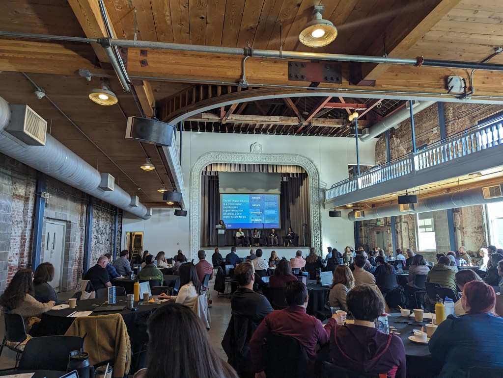 A large industrial room with a crowd of seated people facing a stage with a water workforce presentation
