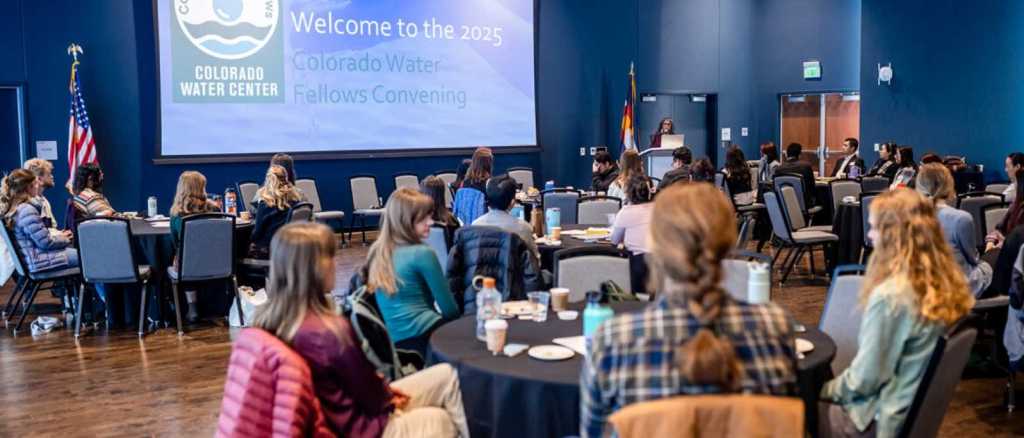 Attendees seated at round tables listen to a speaker at a podium during the 2025 Colorado Water Fellows Convening. A large screen displays the event title and Colorado Water Center logo in a modern conference room with blue walls and wood flooring.