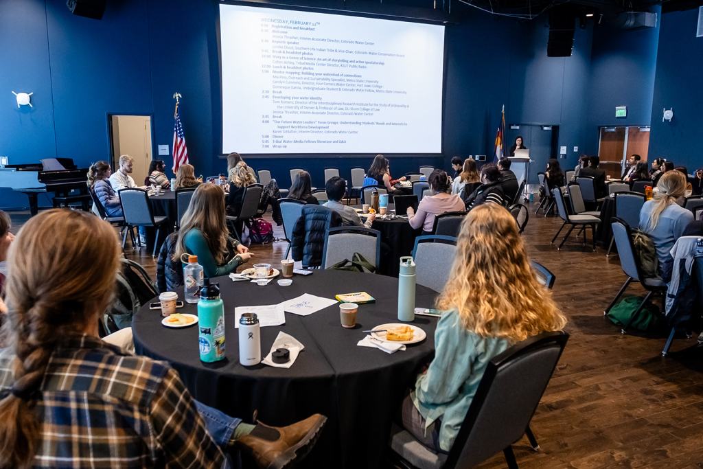 A speaker giving a slideshow presentation addresses a room full of students at round tables from a podium