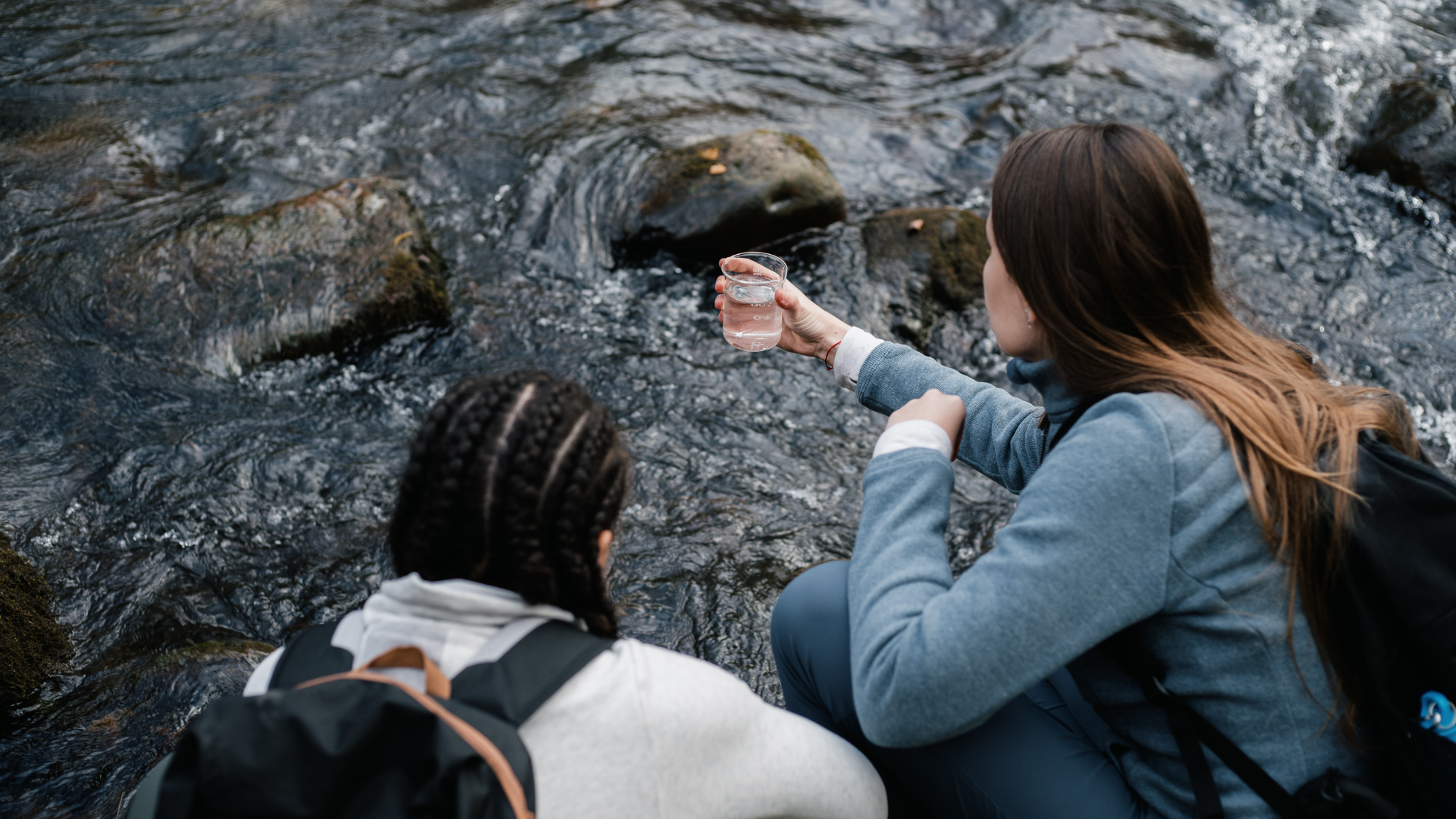 Two people kneeling by a rocky stream, one holding a clear glass of water as if collecting a sample.