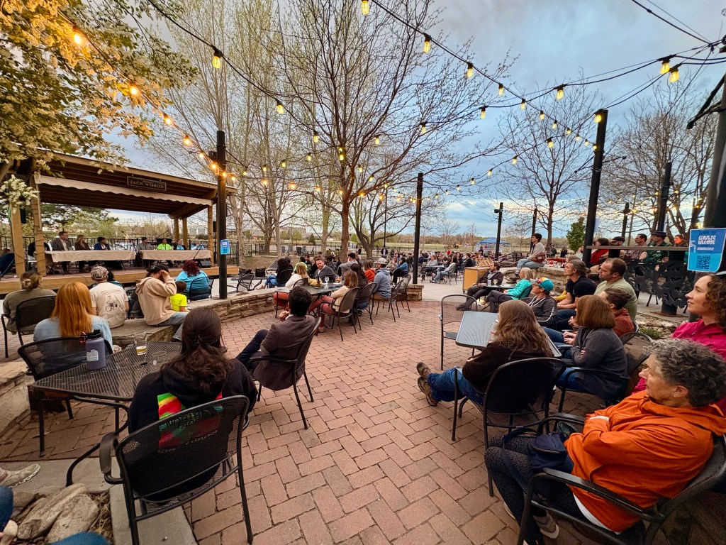 A side view of a crowd of people face a stage with two tables and panelists on an outdoor brewery patio with string lights under a cloudy sky