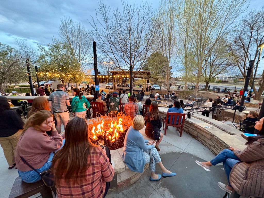 A crowd of people face a stage with two tables and panelists on an outdoor brewery patio with a fire pit in the foreground under a cloudy sky