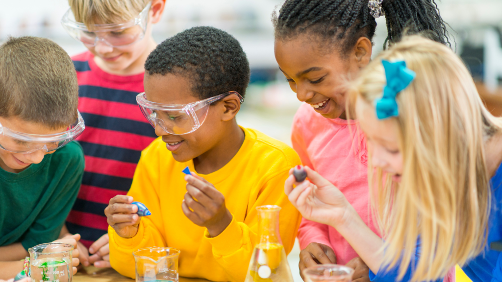 Five elementary school children wear bright colors and science lab safety glasses while putting dye in water beakers for an experiment