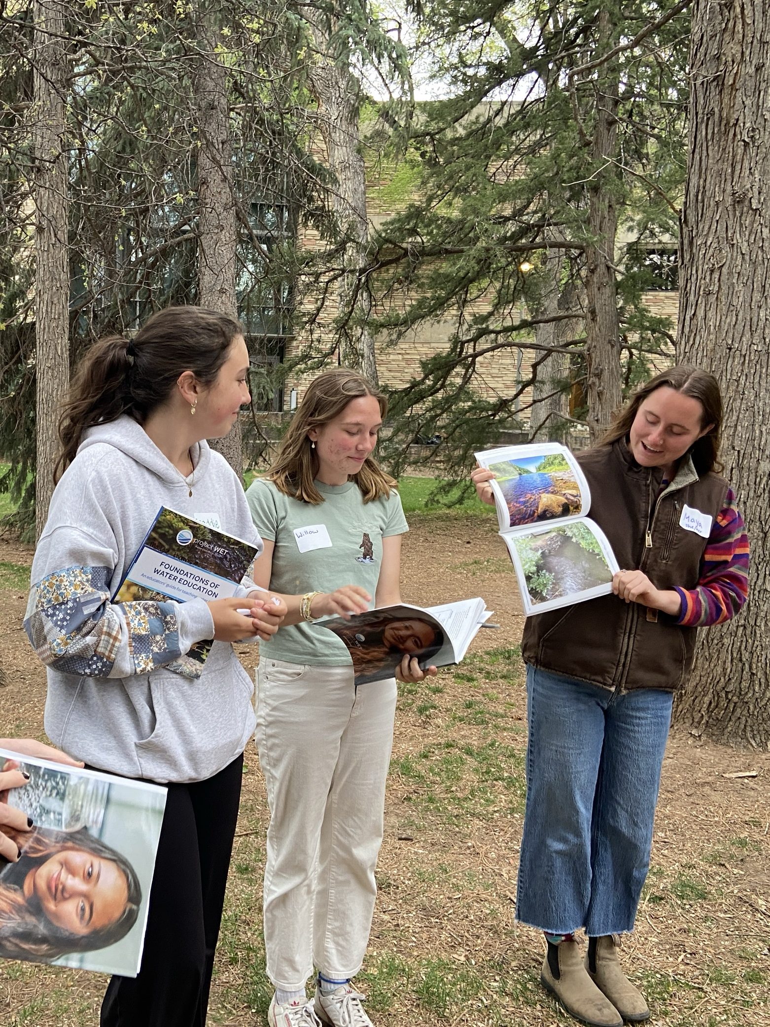 Three women stand outside under tall fir trees holding up science curriculum books