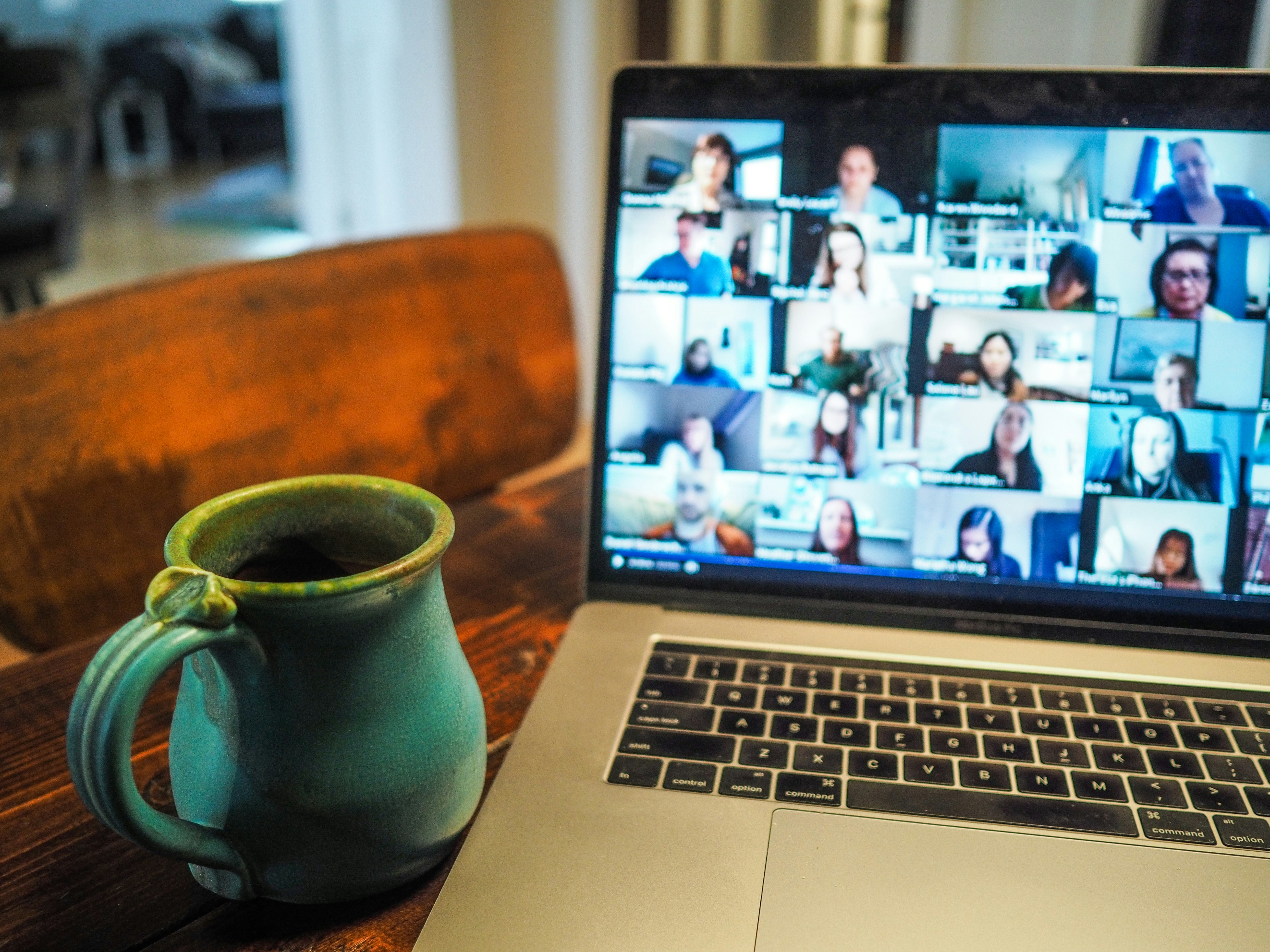 Open laptop displaying a video call with multiple participants in a grid view, next to a teal ceramic mug filled with coffee or tea.