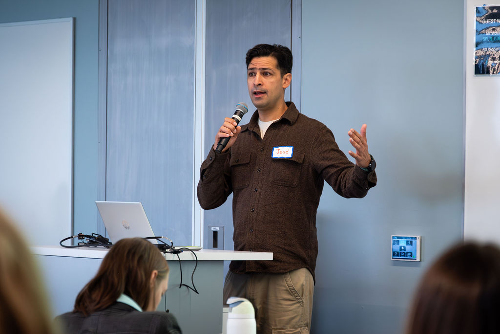A person holding a microphone speaks to an audience while standing at a podium with a laptop, gesturing with one hand during a presentation.