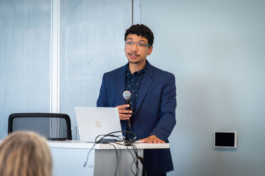 A person in a blue suit and glasses speaks into a microphone while standing at a podium with a laptop during a conference.
