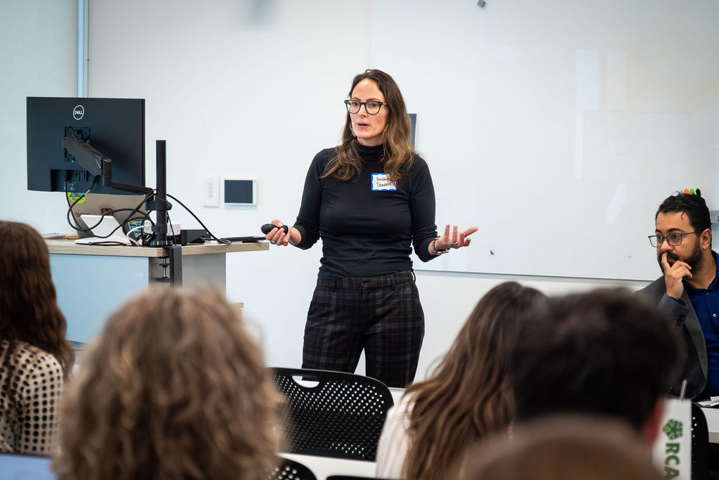 A person stands in front of a seated audience giving a presentation, gesturing with both hands while another person sits nearby.