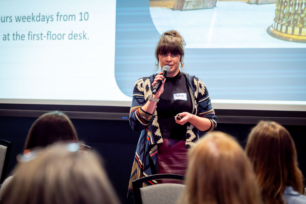 A person holds a microphone and speaks to an audience during a presentation, standing in front of a large projected screen.