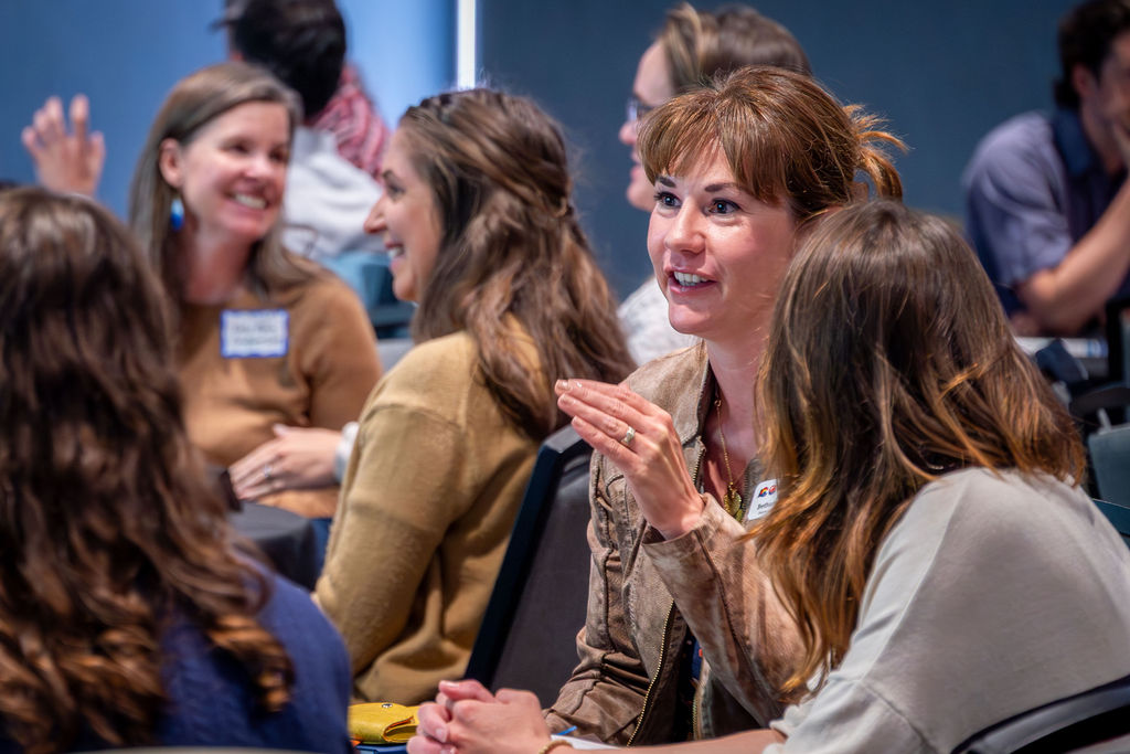 A group of people seated together at a workshop or meeting talk and smile, appearing engaged in conversation.
