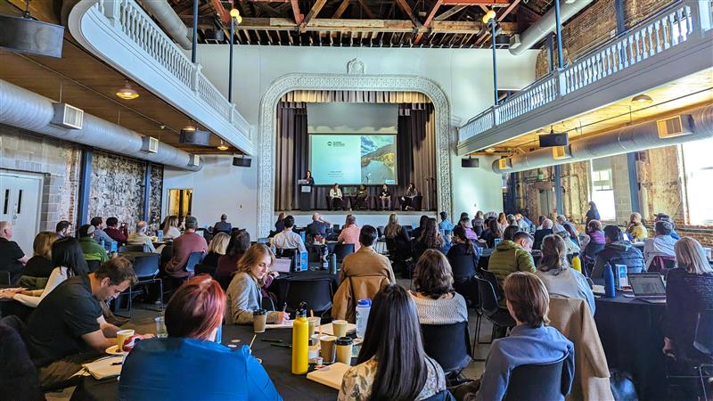A large group of attendees sits at round tables in a historic industrial-style event hall, listening to a panel of speakers on stage beneath a projection screen during a conference session.