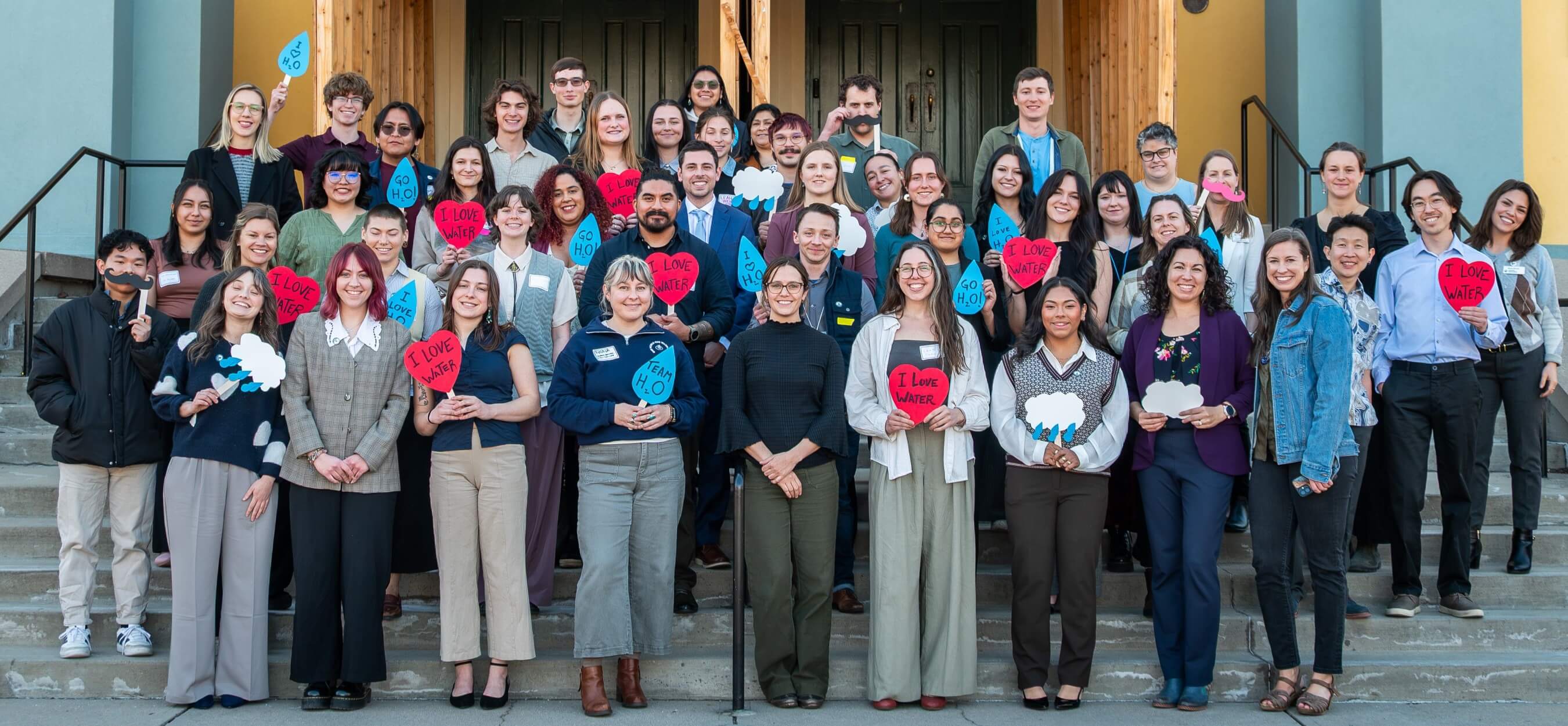 A group photo of Colorado Water Fellows and staff from all six partner institutions, with many people holding red heart signs that say, “I love water,” and other water themed shapes and phrases