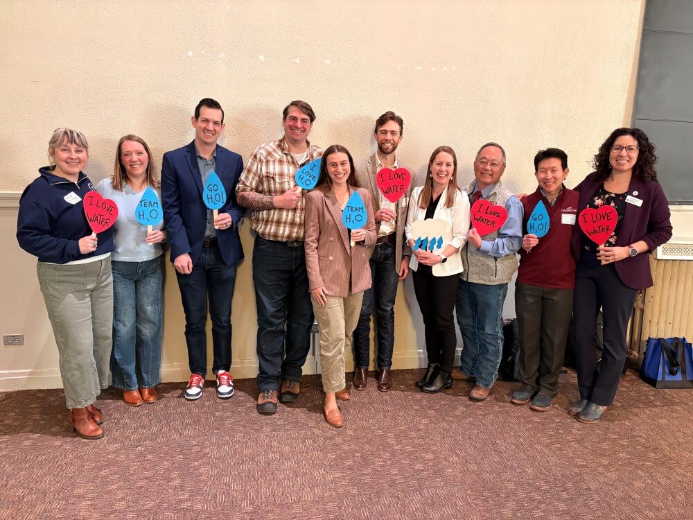 Ten convening speakers, panelists, and moderators standing in front of a wall, holding red heart signs that say “I love water” and other water themed shapes and phrases.
