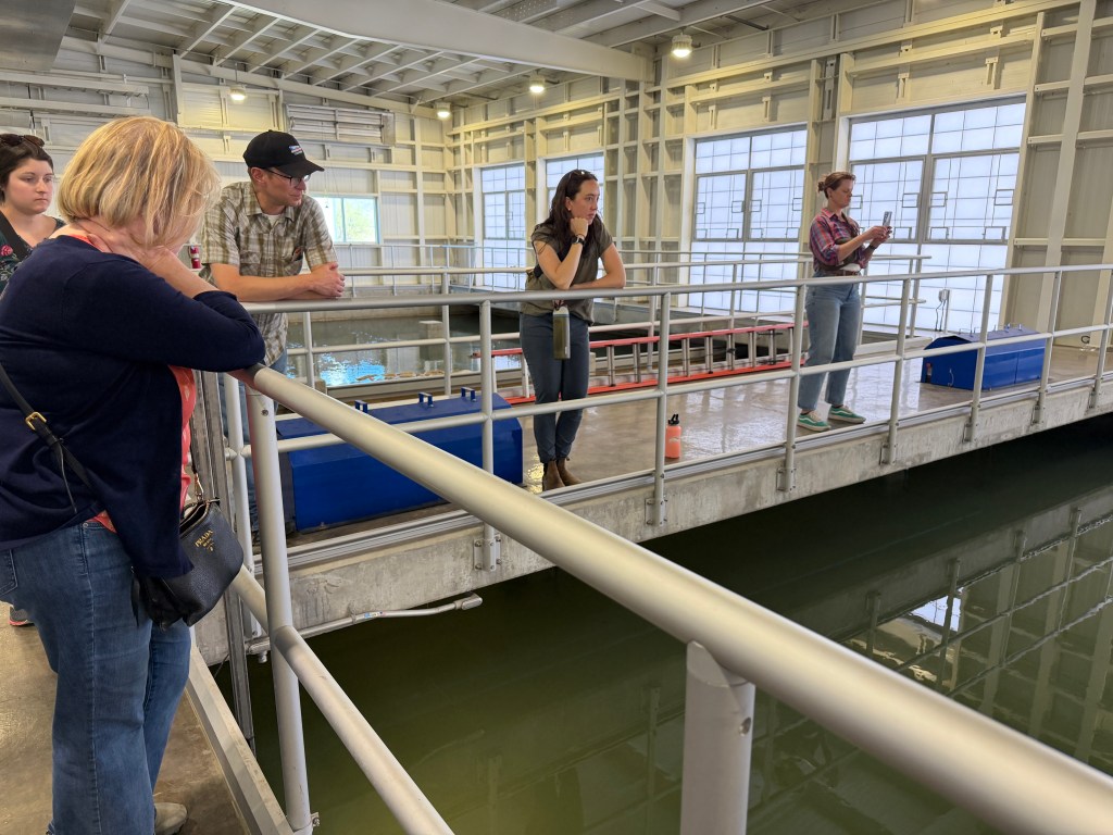 A group of people stands on a platform suspended over water in an indoor facility. Some are looking down at the water while others are listening to someone speaking in the distant.